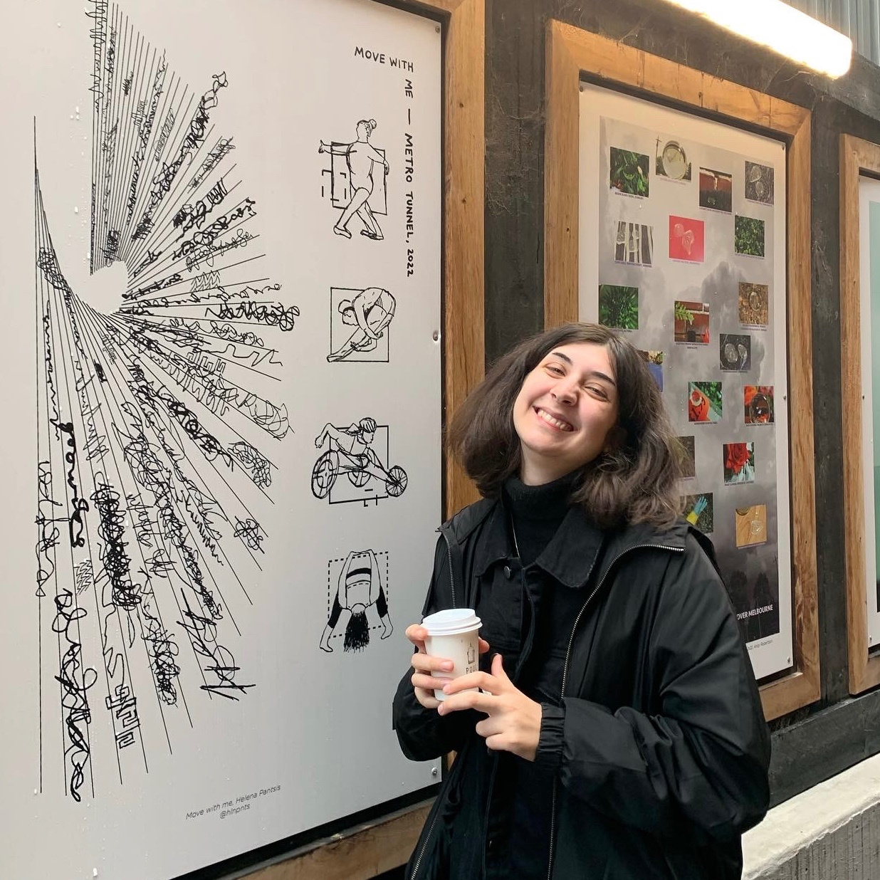 A photo of a young caucasian brunette in a black rain jacket. She holds a coffee cup and stands on front of an abstract monochromatic art piece.